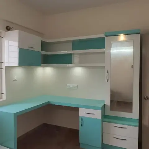 Corner study nook with an L-shaped teal-and-white desk, open display shelving above, and a mirrored wardrobe with an integrated spotlight.