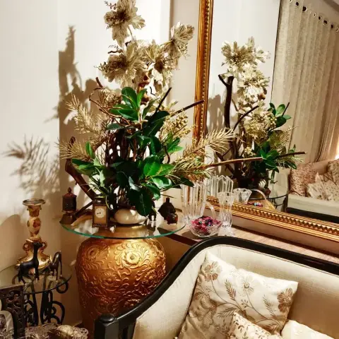 Corner vignette of a carved dark-wood sofa in cream brocade with zari cushions, backed by a tall gilt-framed mirror and a glass top set on an embossed gold urn