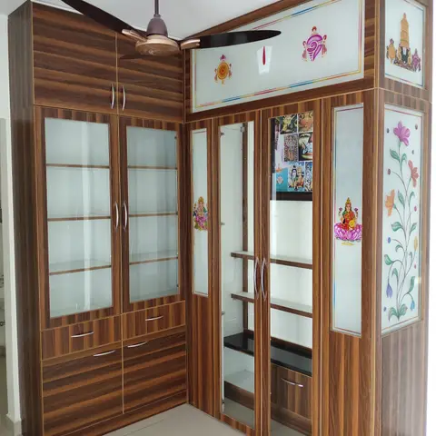 Corner wooden pooja unit with printed deity motifs on frosted glass shutter panels and open display shelving beside it.