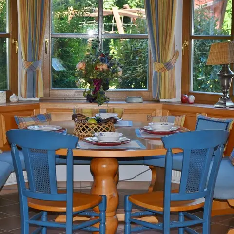 Cottage-style breakfast nook with a round wooden table, built-in blue banquette seating, and striped cushions beside bay windows overlooking a garden.