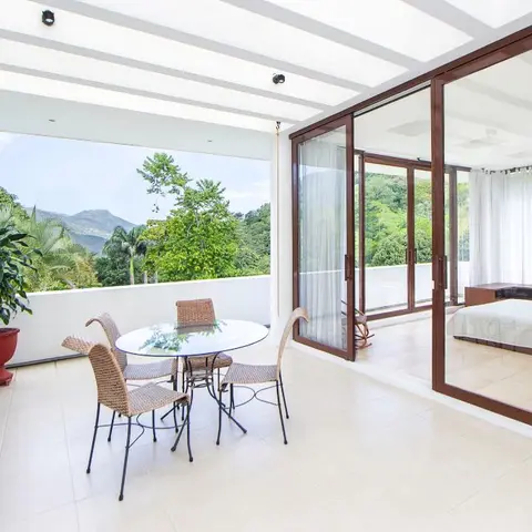 Covered balcony with a round glass table and woven rattan chairs facing a mountain and treeline view, sliding glass doors opening to a bedroom.
