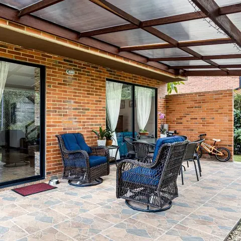 Covered brick-walled home patio with a polycarbonate pergola roof, wicker outdoor armchairs with navy cushions and a matching dining set, opening onto a garden.