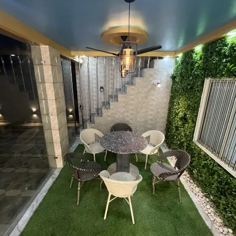 Covered courtyard sit-out at night where six rattan bistro chairs ring a round granite pedestal table on a rectangle of artificial turf edged with white