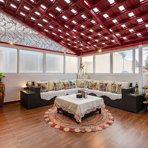 Covered rooftop balcony lounge with a white leaf-pattern jaali screen, a dark rattan corner sofa with patchwork cushions, and a round braided rug.