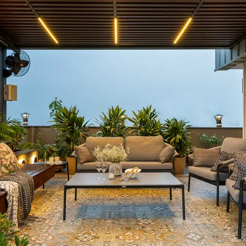 Covered rooftop terrace lounge with patterned cement-tile flooring, grey outdoor sofas, and a slatted pergola with linear pendant lighting at dusk.