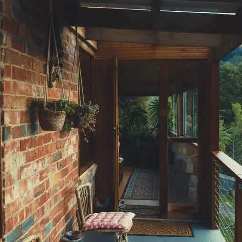Covered timber balcony with an exposed brick wall, hanging macrame plant baskets, a cushioned stool, and a forest valley view beyond the cable railing.