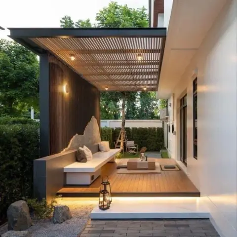 Covered timber deck off a white house at dusk: a slatted pergola with recessed downlights over a built-in white bench against a black batten wall