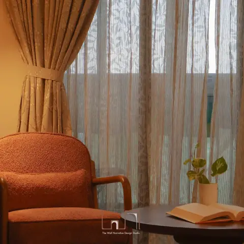 Cozy reading corner with a terracotta boucle armchair beside sheer patterned curtains and a round side table holding an open book and a small potted plant.