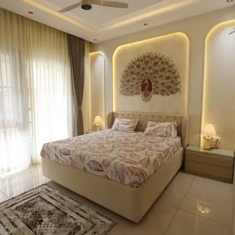 Cream bedroom where a buttoned upholstered bed sits in a backlit arched niche carrying a gold-and-purple metal peacock relief