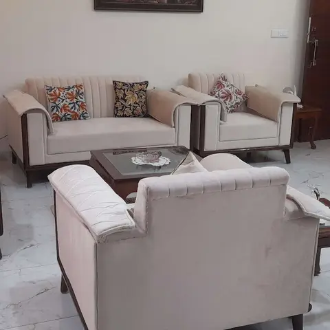 Cream channel-tufted sofa set with floral scatter cushions around a glass-top wooden coffee table on marble flooring, in front of a dark wood main door.