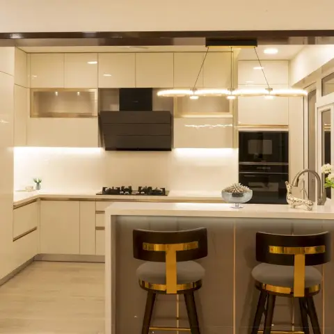 Cream-gloss open kitchen framed by a dark wood portal, with two black-and-gold velvet bar stools pulled up to the quartz breakfast counter under a pair of