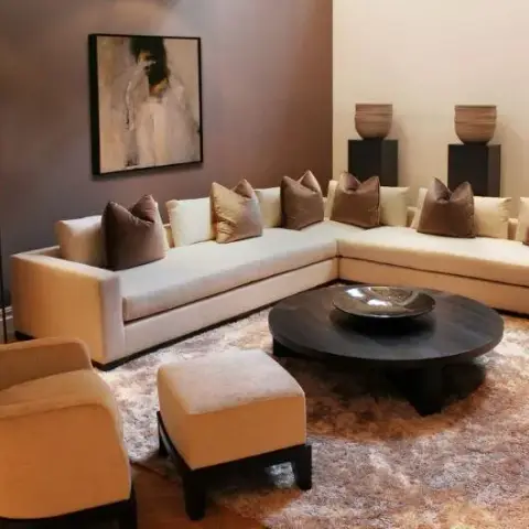 Cream L-shaped low sectional with mocha and gold cushions against a taupe wall, a round ebonised coffee table holding one metal bowl on a shaggy ombre rug
