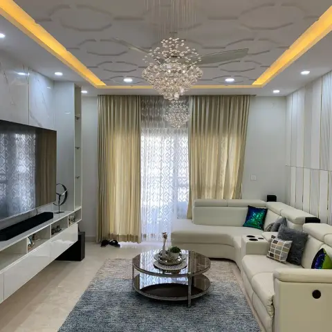 Cream leather recliner sofa and a round smoked-glass table on a blue shag rug, under a honeycomb-relief POP ceiling where a crystal chandelier hangs inside the