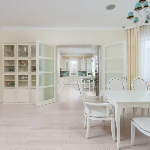 Cream-toned dining room with a glass-front crockery cabinet, French doors opening to the kitchen, and a crystal chandelier over a white dining table.