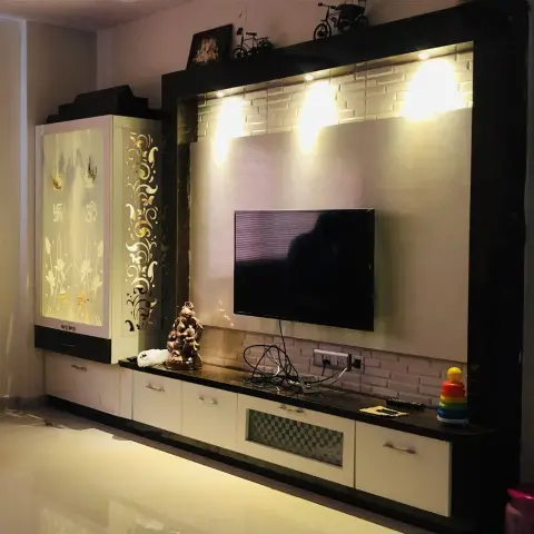 Dark-framed TV wall with white brick-effect cladding lit by three warm spots, sitting next to a backlit pooja cabinet with Om and swastik etched glass and a