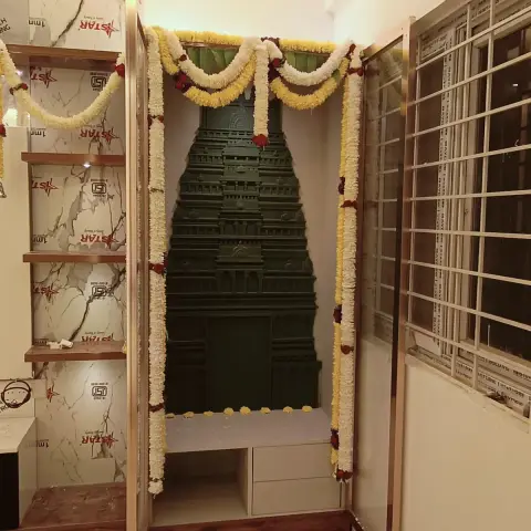 Dark-green carved gopuram mandir seated in a rose-gold framed niche, marigold garlands still draped over the opening and protective film not yet peeled off the