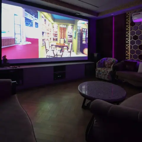 Darkened home theatre room with a wall-filling projection screen over a long low media console, plush taupe sofas and an oval stone coffee table