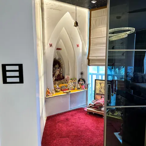 Dedicated pooja alcove with a backlit white arched temple unit, black marble overhead lintel, and a red shag rug, screened off by a glass sliding door.
