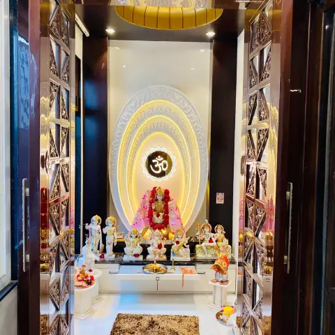 Dedicated pooja room seen through mirror-inlay lattice double doors, with idols on a marble platform against a concentric carved oval backdrop lit warm behind