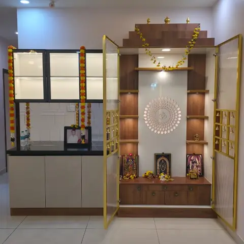 Dedicated pooja room with a tiered wooden mandir unit, a backlit Om medallion niche, gold-trimmed glass doors hung with bells