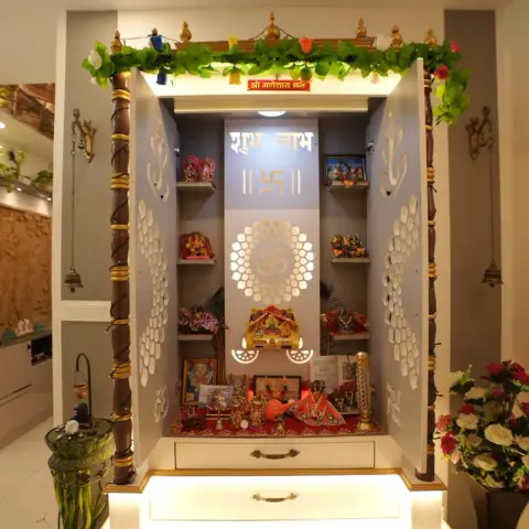 Dedicated pooja unit with carved white jaali cabinet doors, gold-trimmed pillars, a floral garland, and idols arranged on illuminated shelves.