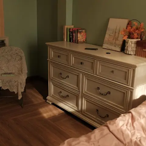 Dim bedroom corner where a cream moulded chest of drawers holding paperbacks, dried orange leaves and a wicker vanity case stands against a sage wall