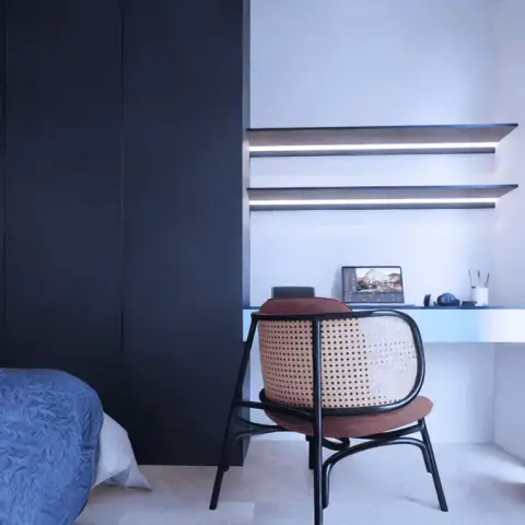 Dim bedroom render looking past the corner of a bed with a blue cable-knit throw to a navy wardrobe wall and a white work niche with two LED-lit floating
