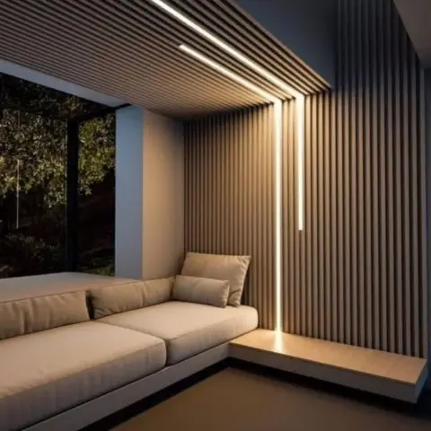 Dim seating nook where a fluted oak slat wall wraps onto a slatted ceiling raft and a single recessed light line runs unbroken from ceiling to floor at the