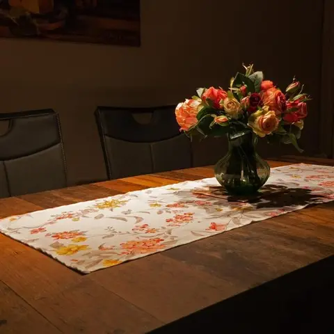 Dimly lit dining table in reclaimed wood with black leather-back chairs, a floral table runner, and a glass vase of orange roses as the centerpiece.