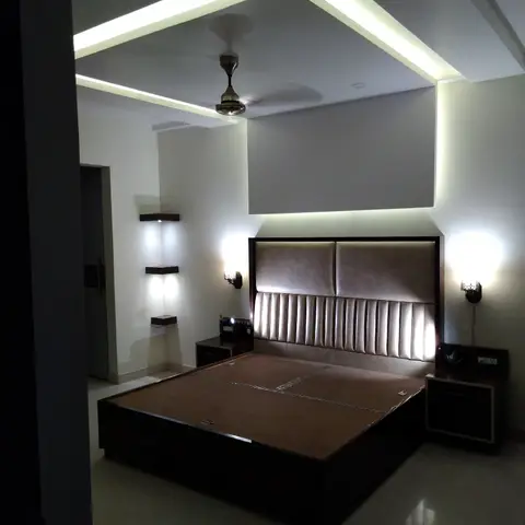 Dimly lit master bedroom with a channel-tufted beige headboard bed and wall sconces either side, under a box-shaped cove-lit false ceiling frame.