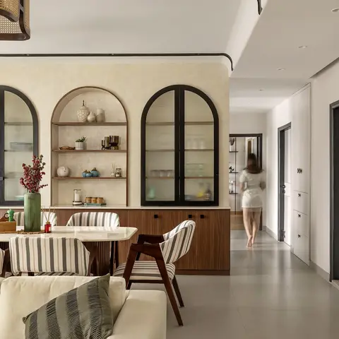 Dining and living area with arched glass-front display cabinets and open shelving on a plastered wall, a marble-top dining table with striped chairs
