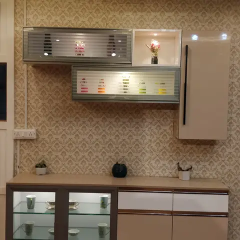 Dining-area crockery unit with backlit glass display shelves holding cups and bowls, a damask wallpaper backdrop, and beige cabinetry with a wood countertop.