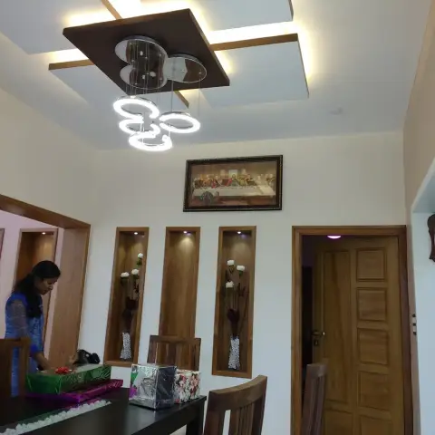 Dining area of a Kerala home with three teak-framed lit wall niches holding white flower vases below a framed Last Supper print