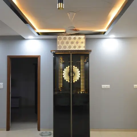 Dining area view of a floor-to-ceiling backlit black-glass pooja unit with a lotus-motif jaali screen, set beneath a stepped cove-lit false ceiling.
