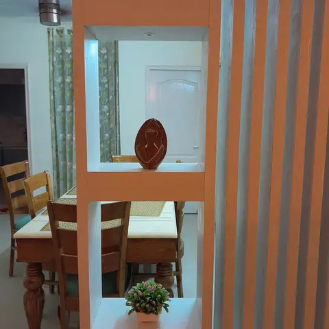 Dining area with a carved wood table and chairs seen through an open shelf partition displaying a Buddha head and a carved Ganesh idol.