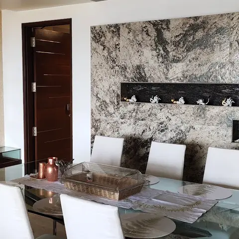 Dining area with a glass table and white leatherette chairs against a textured stone feature wall with lit display niches and a family photo gallery