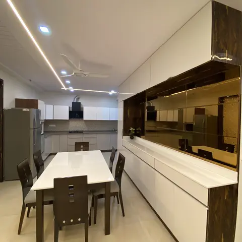 Dining area with a glossy white table and dark wood chairs beside a long crockery unit finished in bronze mirror and a jaali-cutout end panel