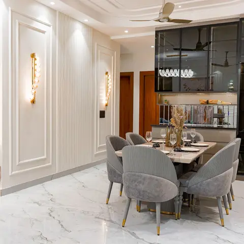 Dining area with a round marble-top table and six channel-tufted grey velvet chairs, gold wall sconces, a ribbed panel wall