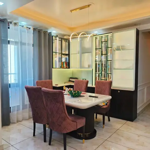 Dining area with a round marble-top table, mauve quilted velvet chairs on brass legs, a backlit glass crockery cabinet