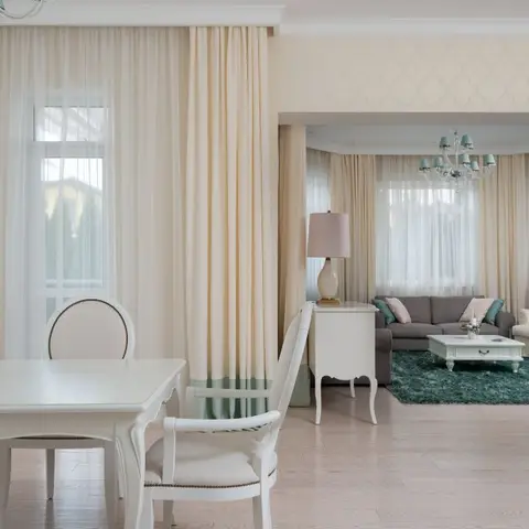 Dining area with a white cabriole-leg table and upholstered chairs, floor-length cream curtains, opening into a living room with a teal shag rug beyond.