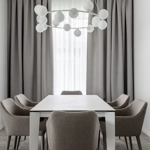 Dining area with a white table and grey upholstered chairs beneath a spherical bubble chandelier, floor-to-ceiling grey curtains