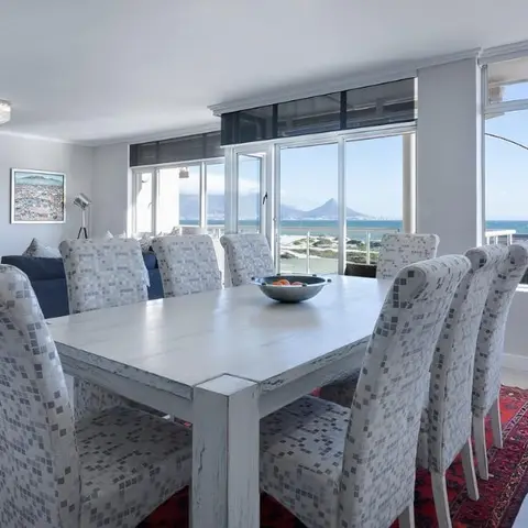 Dining area with a whitewashed wood table and mosaic-patterned upholstered chairs on a red patterned rug, sea view through floor-to-ceiling windows.