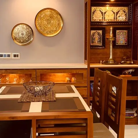 Dining area with a wood table, brass decorative wall plates, and an adjoining built-in pooja unit with Tanjore-style deity paintings.