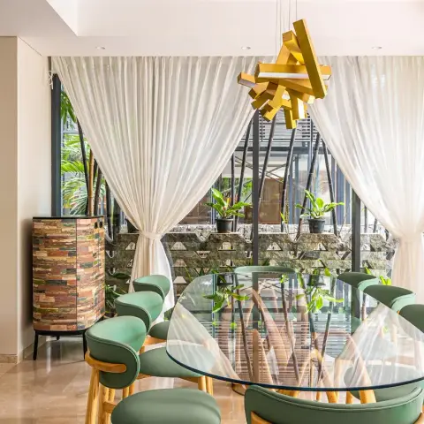 Dining area with an oval glass table on a branch-form timber base and sage green leather chairs, under a sculptural mustard-yellow pendant