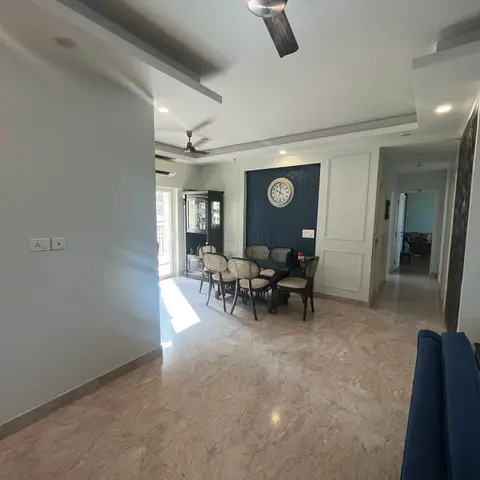 Dining area with cane-back chairs around a glass table beside a navy accent wall with white panel moulding and a round wall clock, opening to a hallway