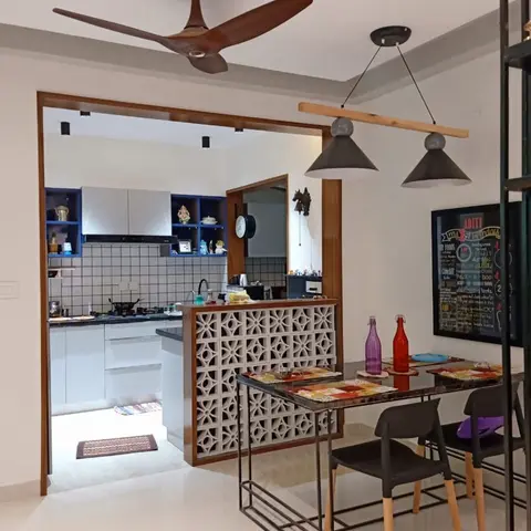 Dining nook divided from the kitchen by a decorative concrete jaali screen, lit by twin cone pendant lights over a glass-top table.