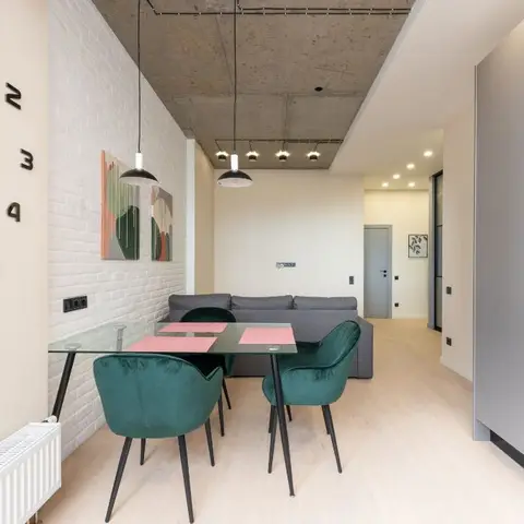 Dining nook with a glass table and emerald velvet chairs against a white exposed-brick wall, black pendant lights hanging from a raw concrete ceiling