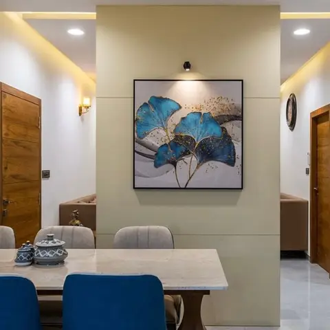 Dining nook with a marble-top table, royal-blue velvet chairs, and a spotlit botanical canvas on a partition wall beside a wood-framed doorway.