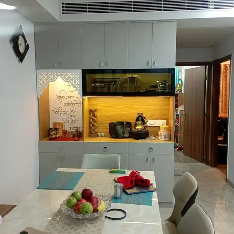 Dining nook with a marble-top table, upholstered chairs and a glass-front crockery unit with a backlit pooja niche built into the same wall.