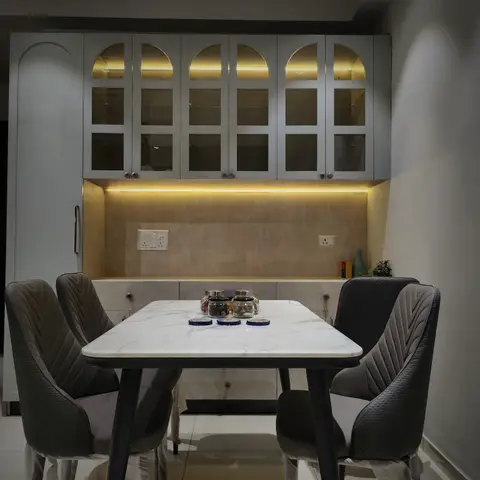 Dining nook with a marble-top table and grey channel-tufted chairs beneath an arched-glass crockery unit lit with warm cove lighting.